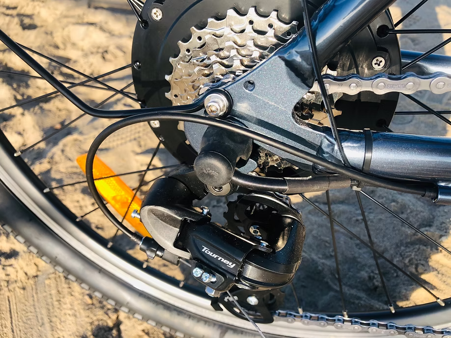 Close-up of a bicycle rear derailleur and cassette, with the brand Tourney visible on the black derailleur component. The bike frame is dark gray, and the background shows sandy ground outdoors.