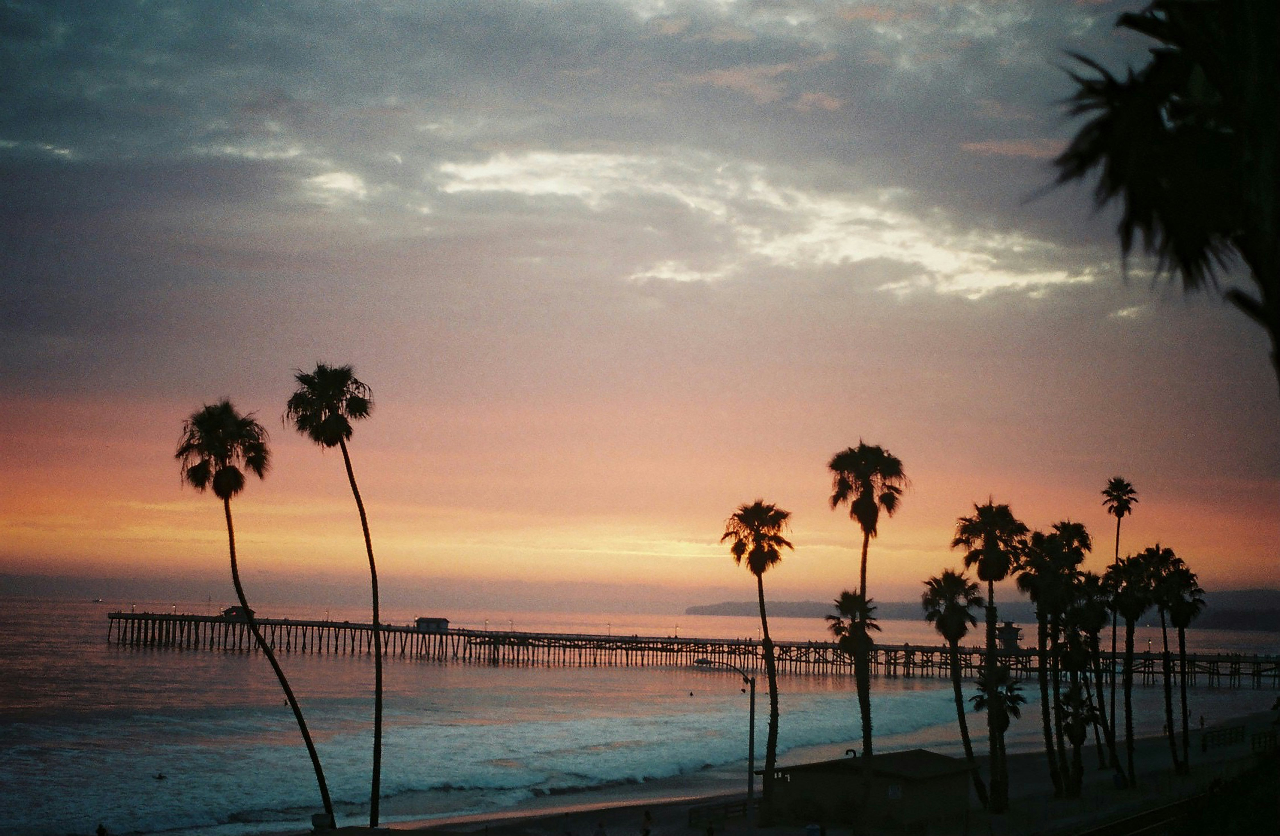 A scenic sunset beach landscape with silhouetted palm trees, a long pier extending into the ocean, and a sky transitioning from warm orange and pink near the horizon to darker blue-gray clouds above.